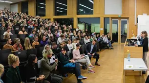 A photo of the front part of the lecture hall taken from the side. David Frum and Prof. Dr. Gerhardt are standing behind the lectern. Parts of the audience are visible. A male student is in the process of asking a question.